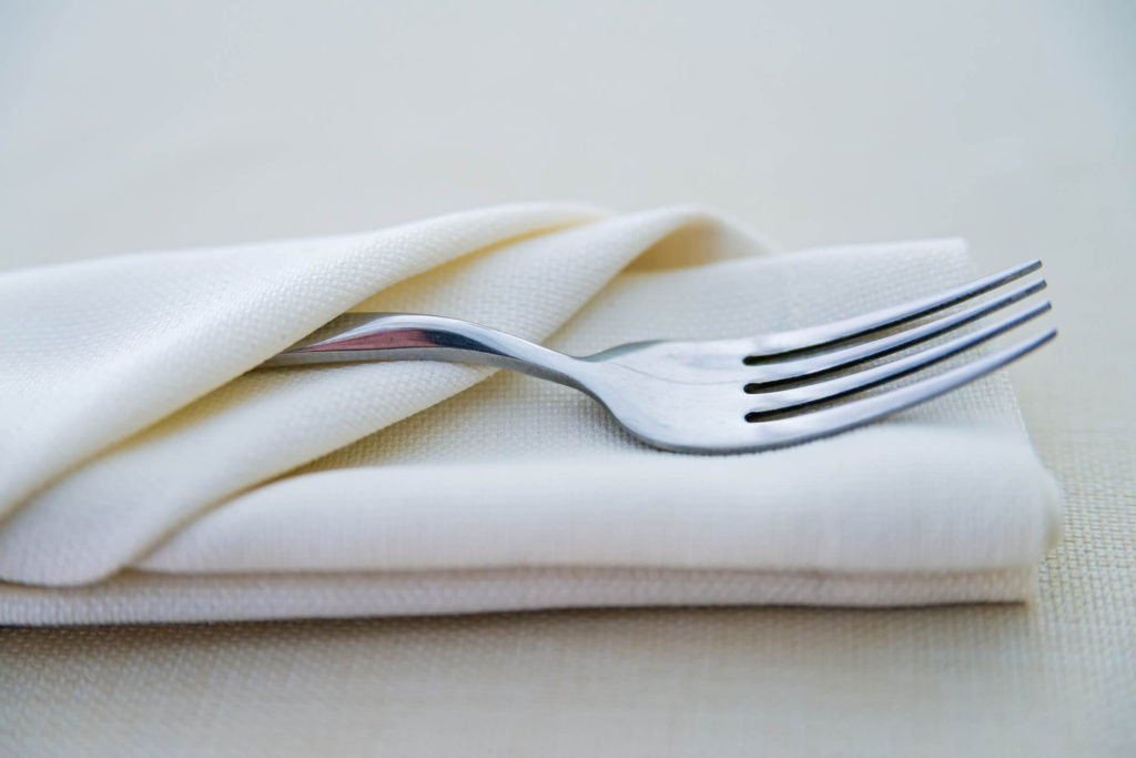 Fork wrapped in a commercial table linen napkin on cream colored tablecloth.