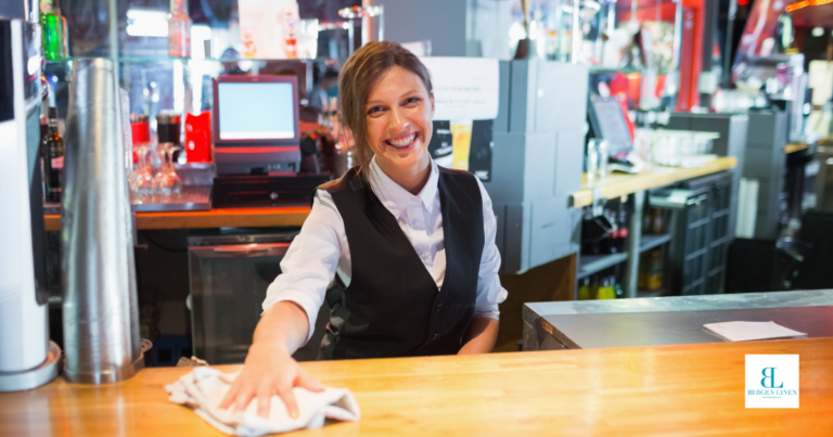 Female bartender in white button-down and black vest wiping down a wood bar counter top with a white bar mop towel.