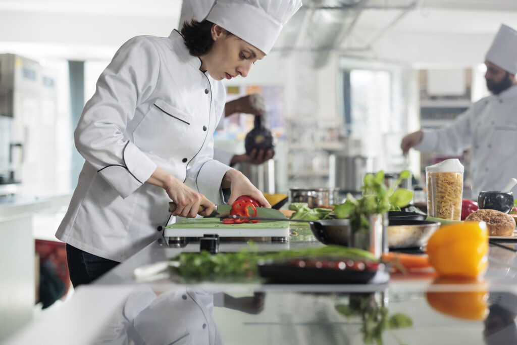 Restaurant commercial kitchen with a sous chef in the forefront chopping fresh vegetables. Other team members in the background.